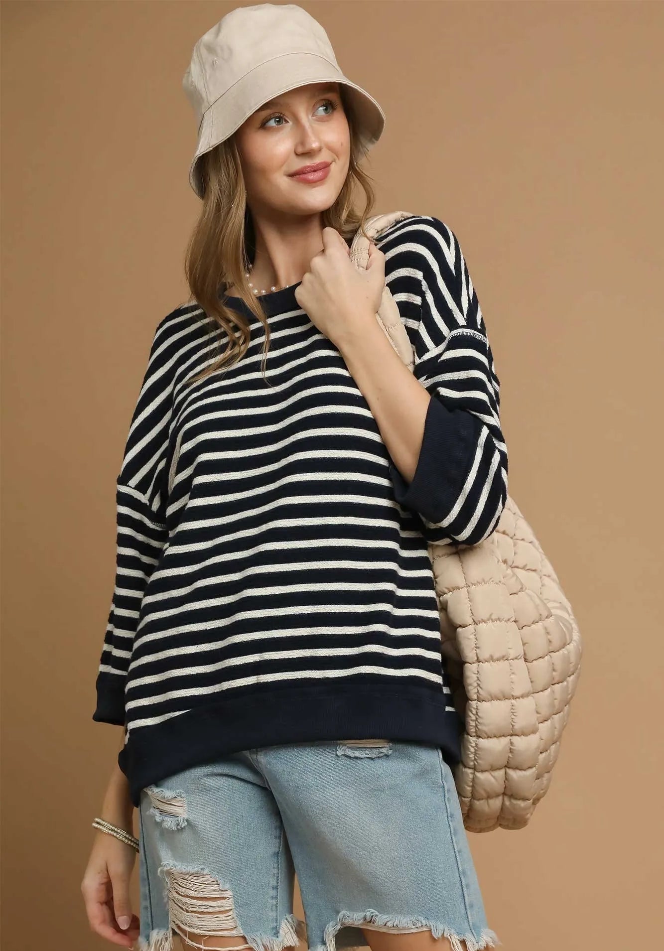 Woman wearing a striped sweater, denim shorts, and a beige bucket hat against a brown background.