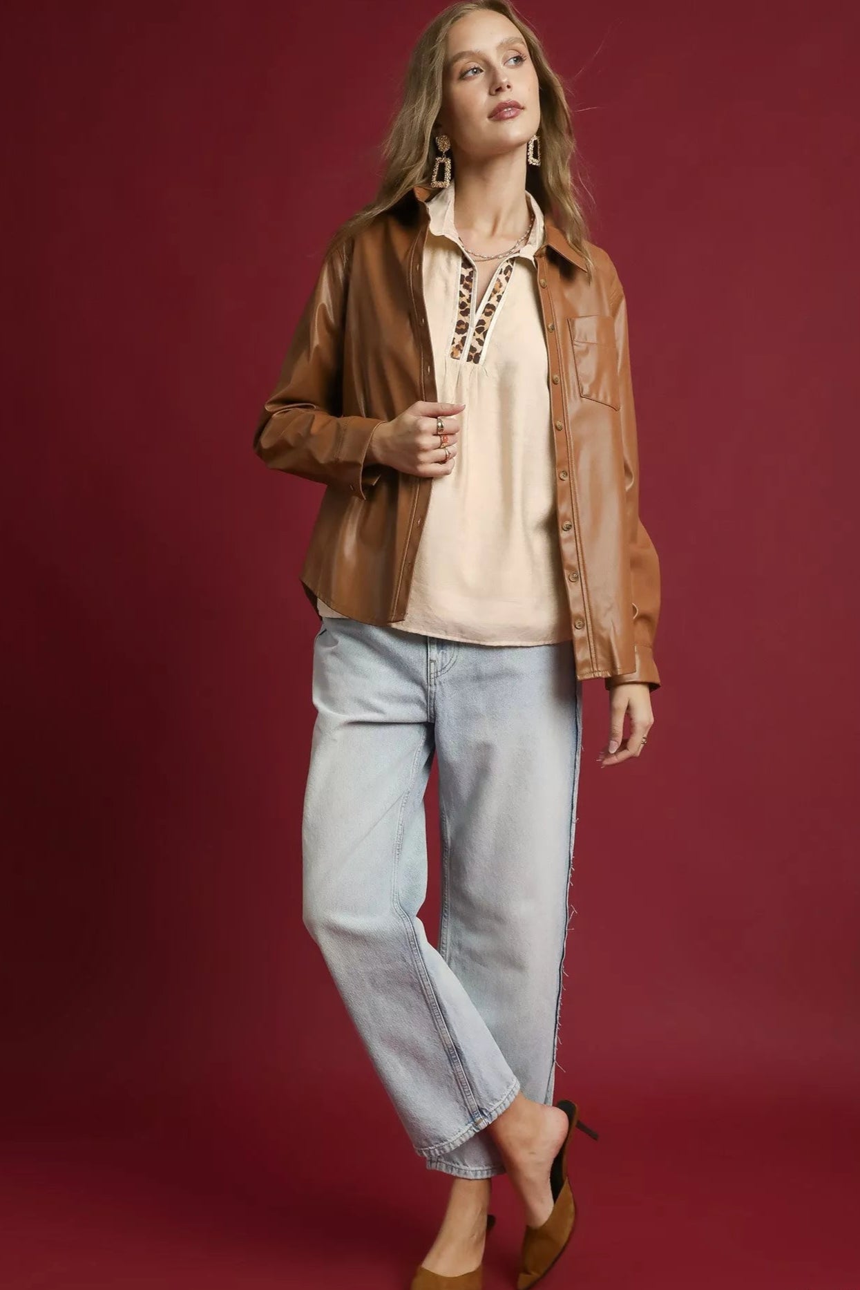 Woman wearing a brown jacket, beige top, and light blue jeans on a red background