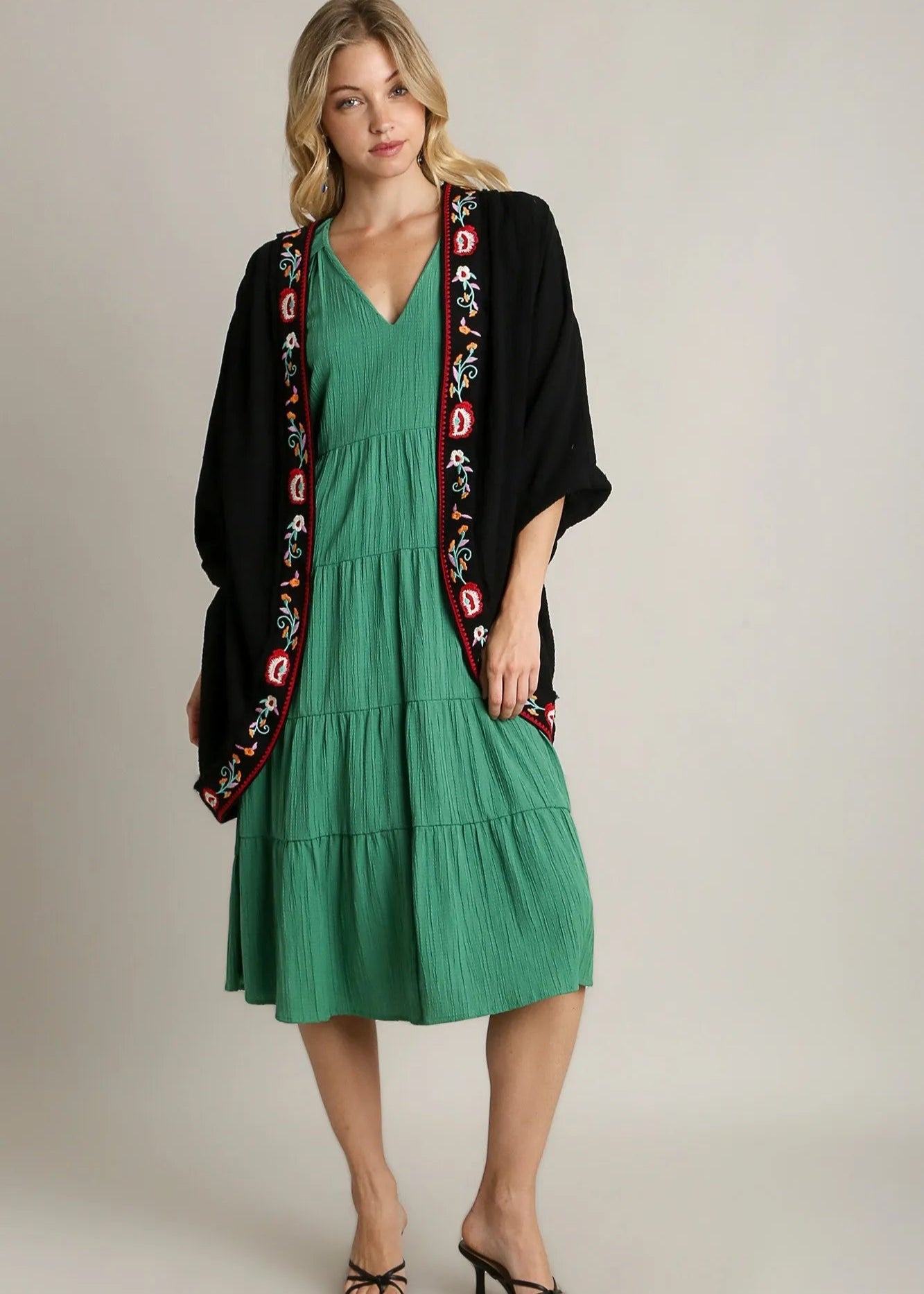 Woman wearing a green dress with a black cardigan featuring colorful embroidery on a plain background