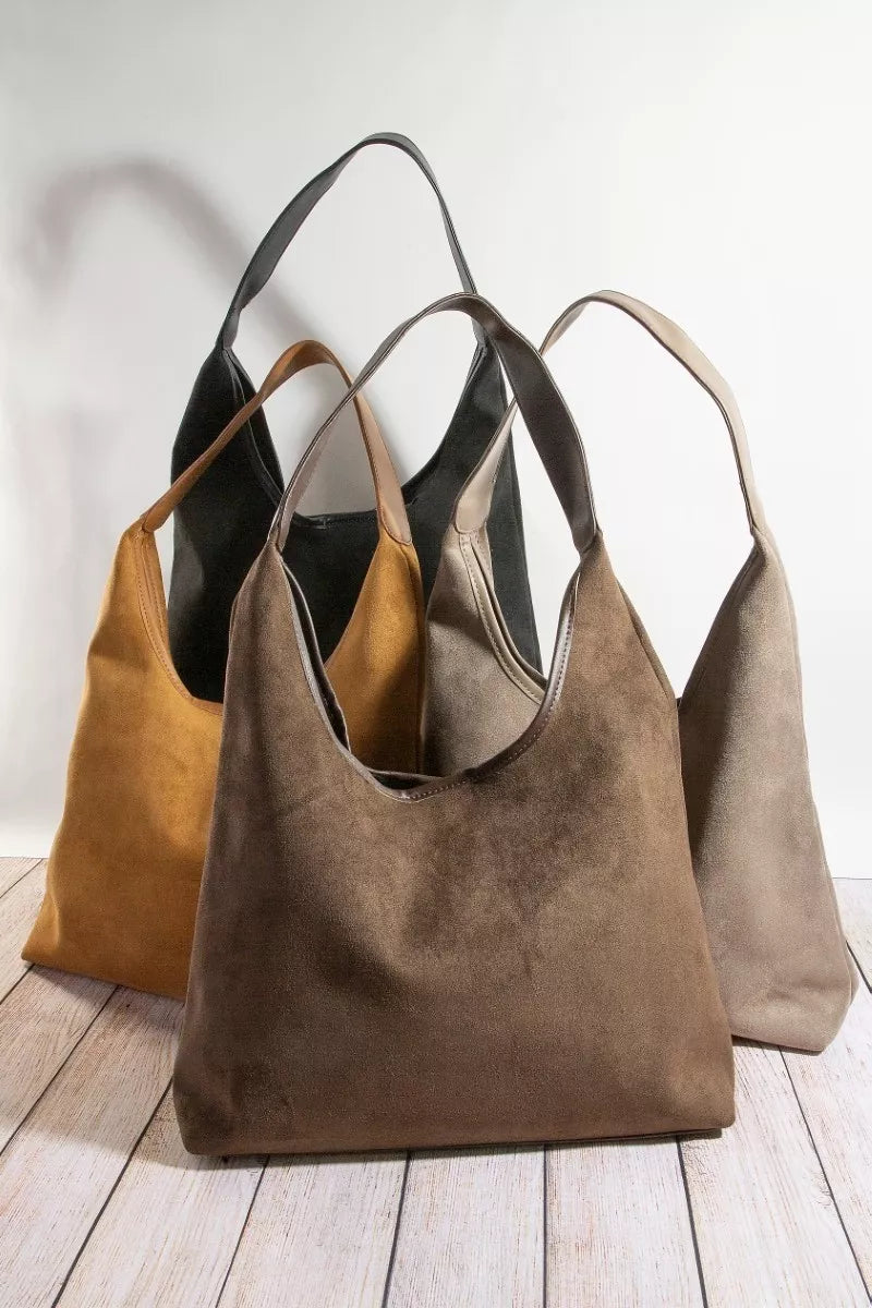 Three suede handbags in brown, black, and beige on a wooden surface with a white background