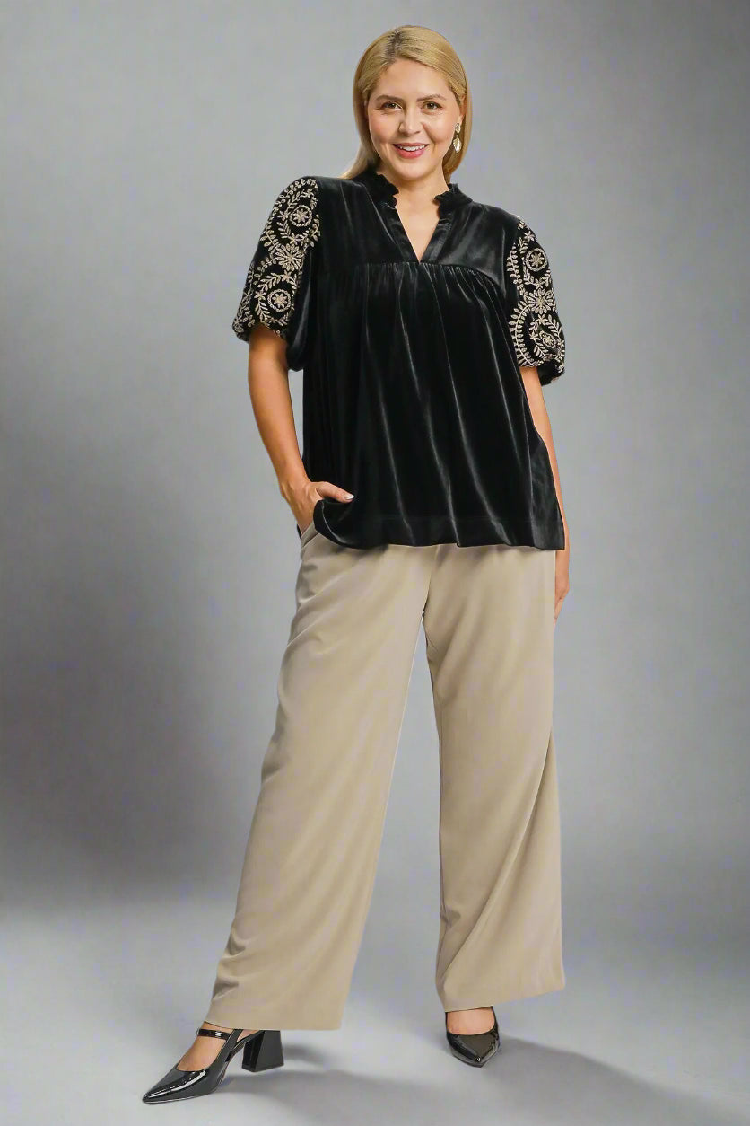 Woman wearing a black velvet blouse with gold embroidery and beige pants on a white background