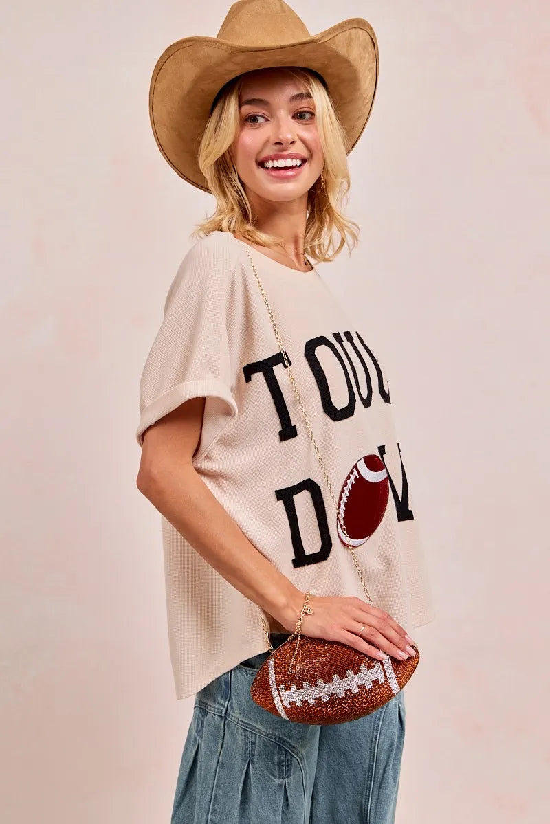 Woman wearing a beige t-shirt with 'Touchdown' text and a football graphic, holding a football, on a light pink background.