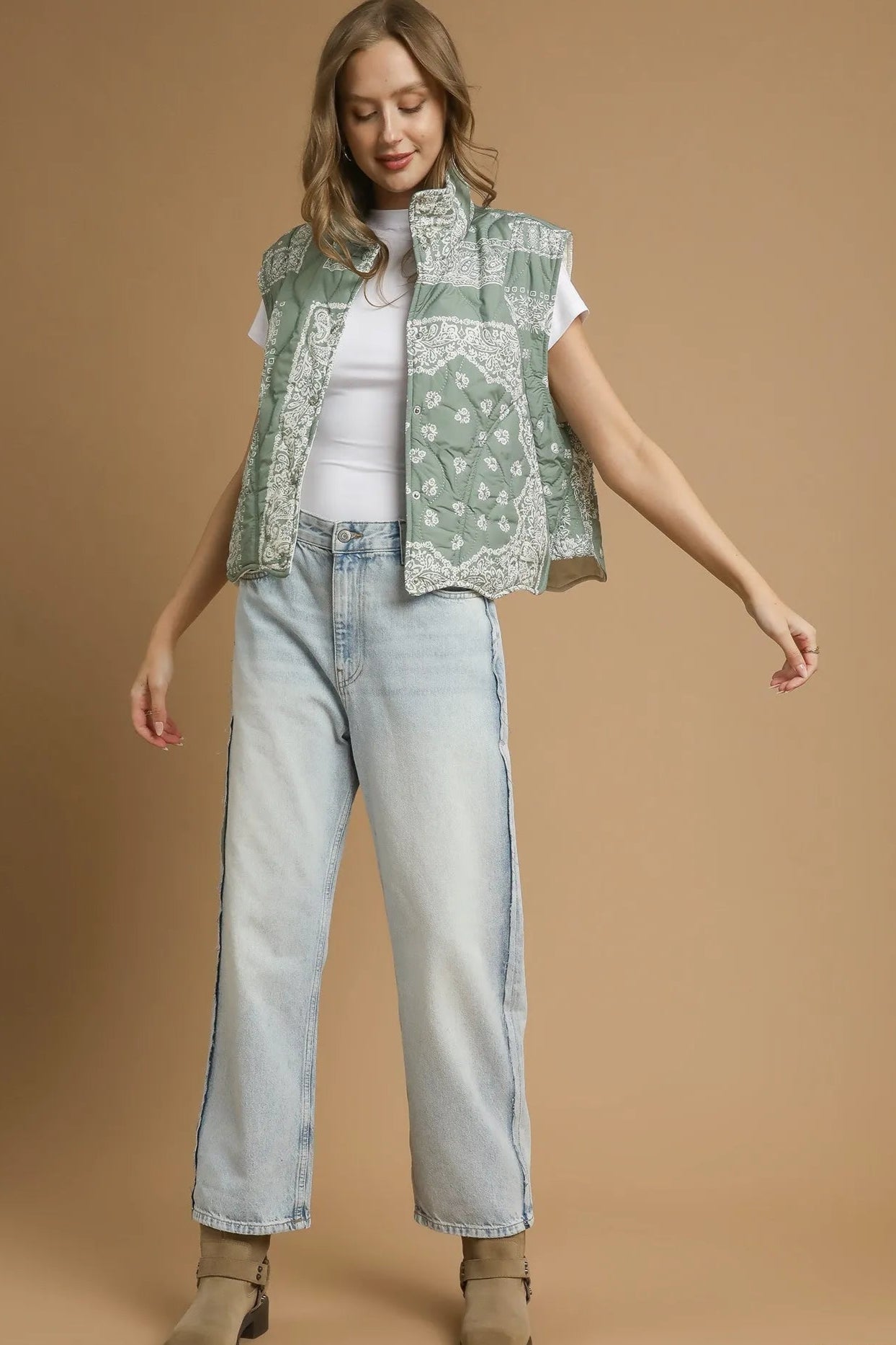 Woman wearing a green patterned vest, white shirt, and light blue jeans on a brown background