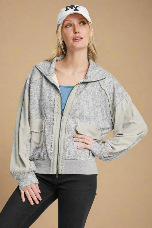 Woman wearing a gray zip-up jacket, black pants, and a white cap on a light beige background