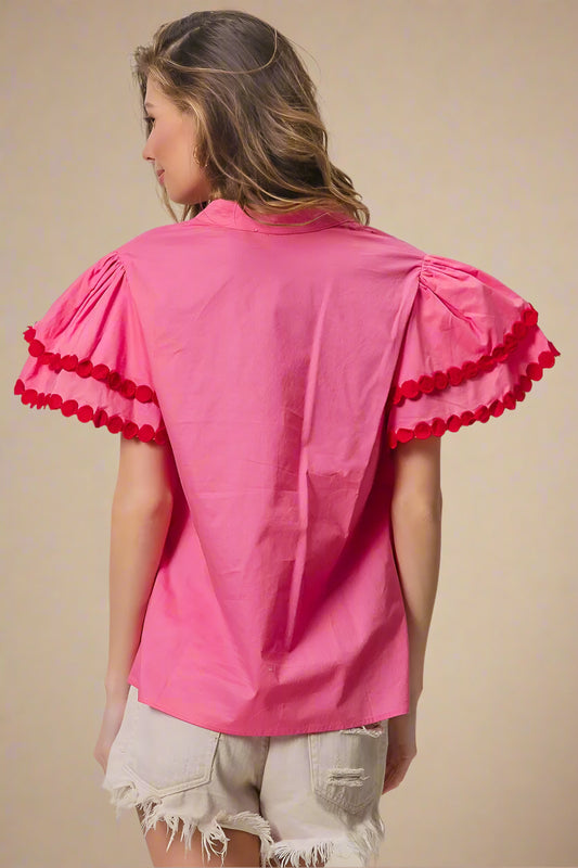 Pink blouse with red pom-pom trim worn by a person on a beige background