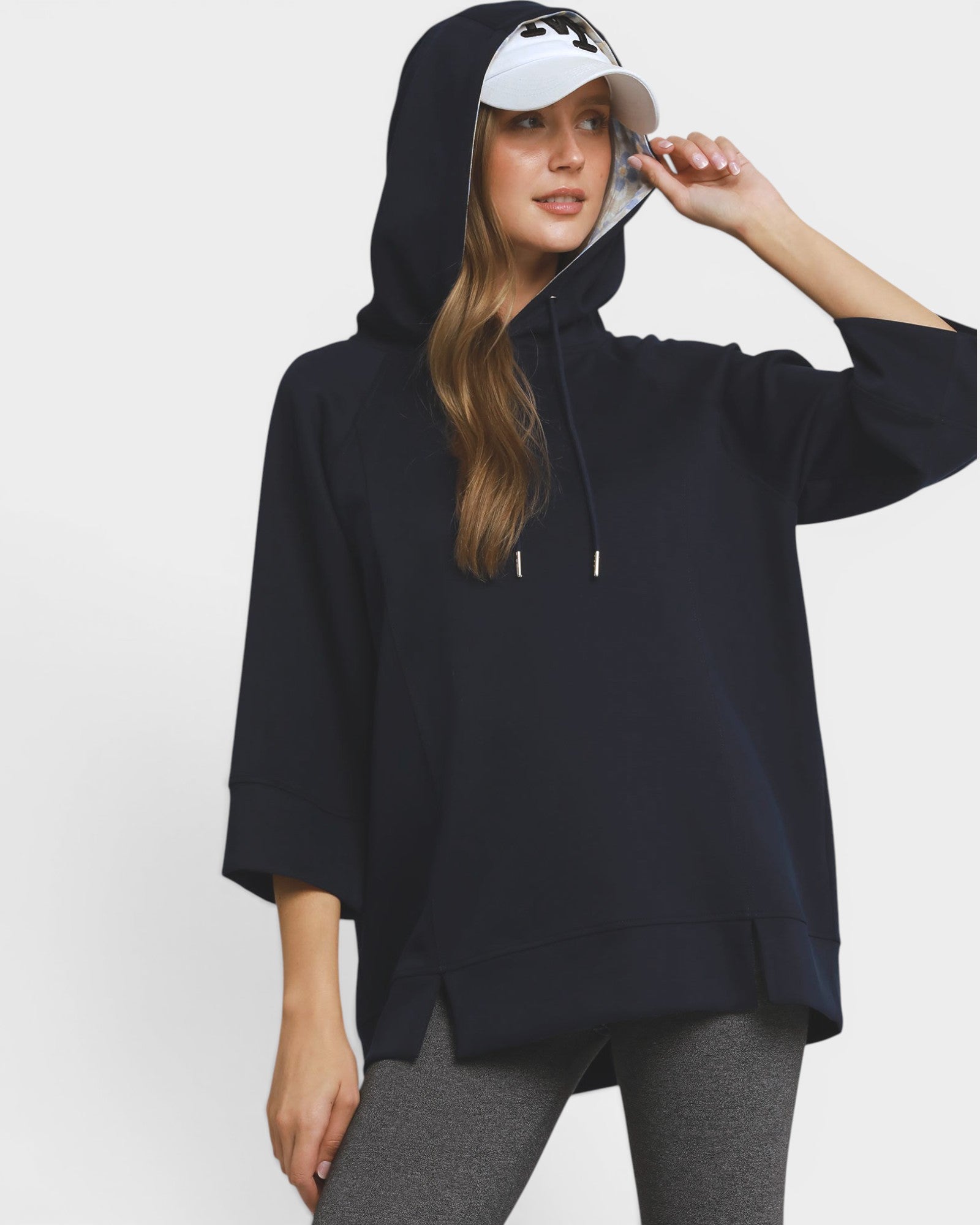 A woman wearing an oversized navy pullover hoodie with three-quarter sleeves, a drawstring hood, and a side slit hem, paired with gray leggings and a white visor, against a plain light gray studio background.
