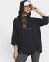 A woman wearing an oversized navy pullover hoodie with three-quarter sleeves, a drawstring hood, and a side slit hem, paired with gray leggings and a white visor, against a plain light gray studio background.