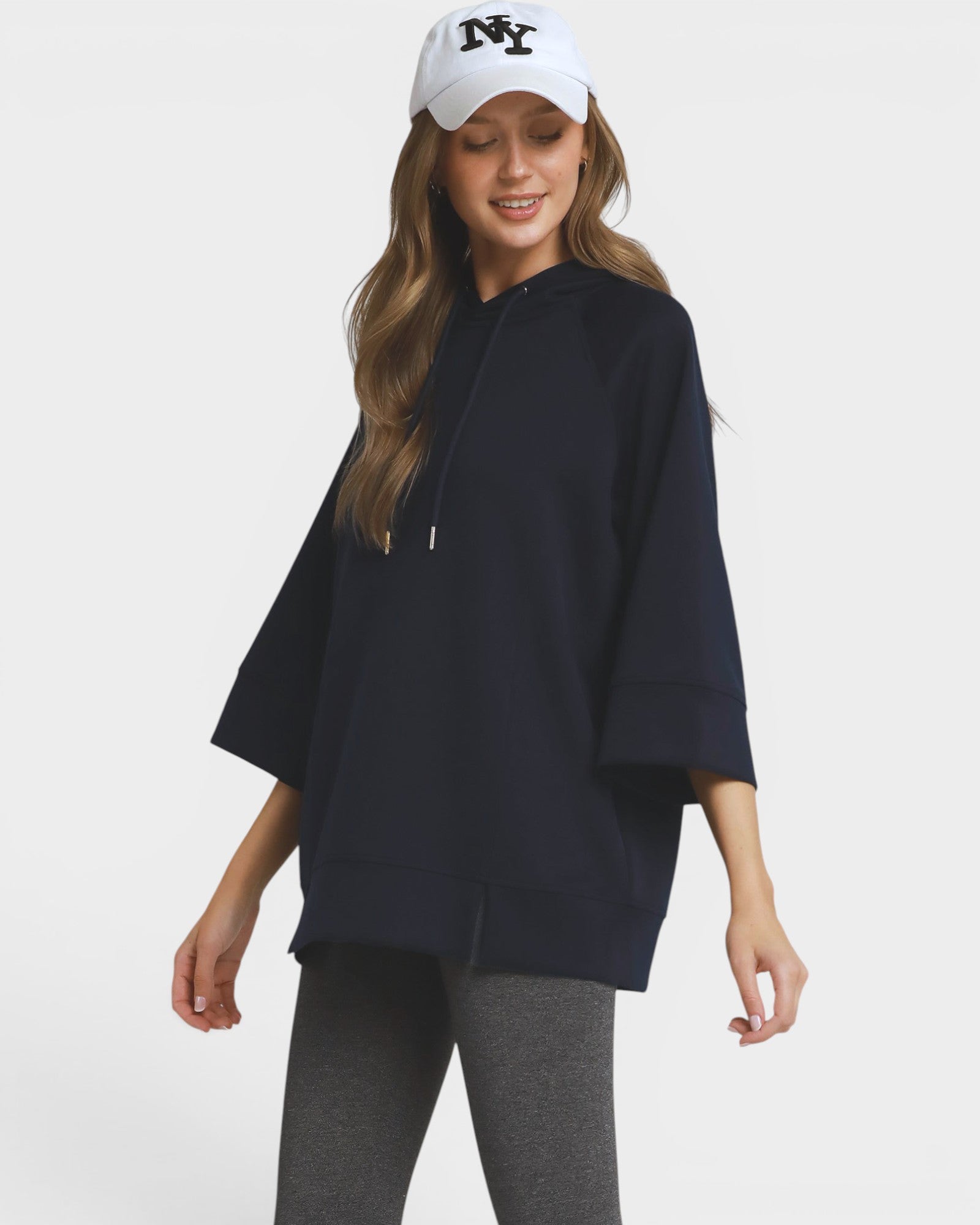 A woman wearing a navy blue oversized pullover hoodie with wide three-quarter sleeves, paired with gray leggings and a white baseball cap with a black 'NY' logo, set against a plain light gray background.
