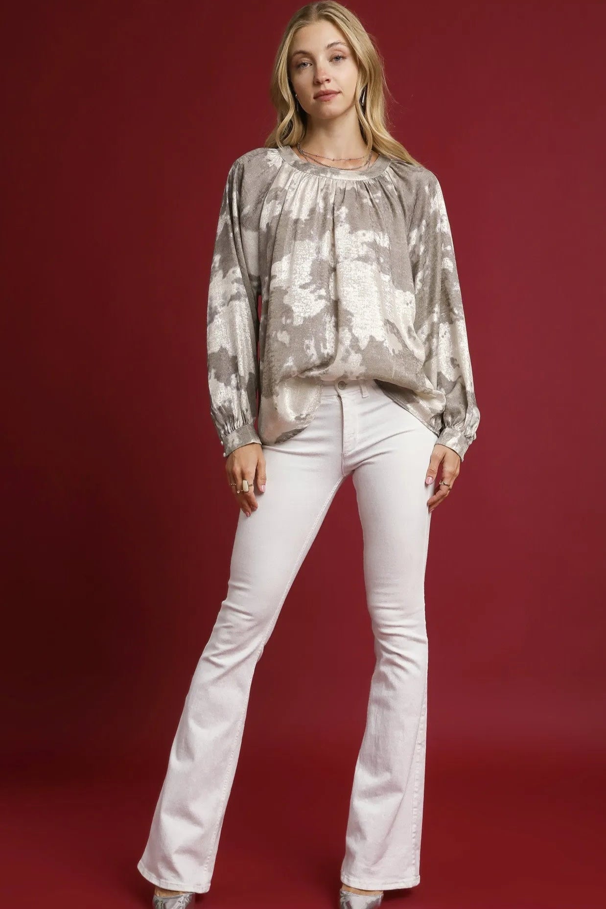 Woman wearing a taupe patterned top and white pants on a red background