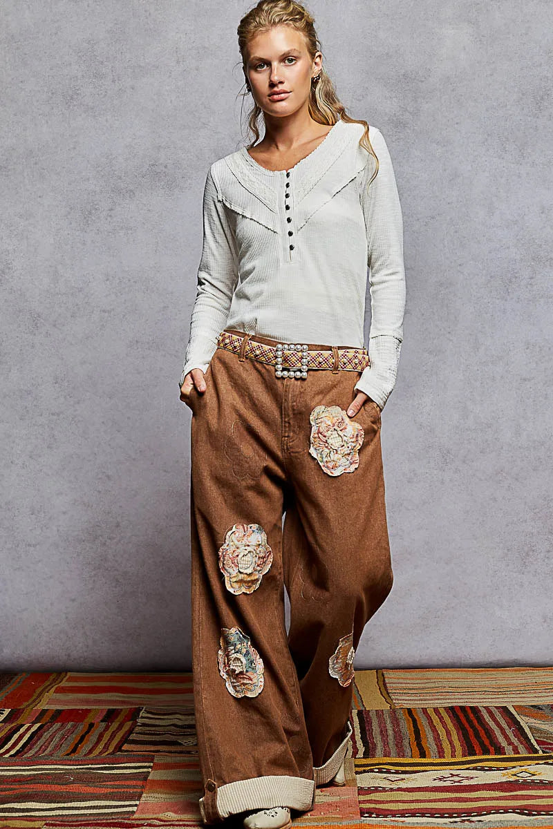 Woman wearing a light gray top and brown pants with floral patches against a gray background