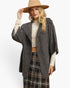 A woman modeling a dark gray textured open-front poncho-style cardigan over a cream mock-neck top and a black plaid skirt, wearing a tan wide-brim hat against a plain white studio background.
