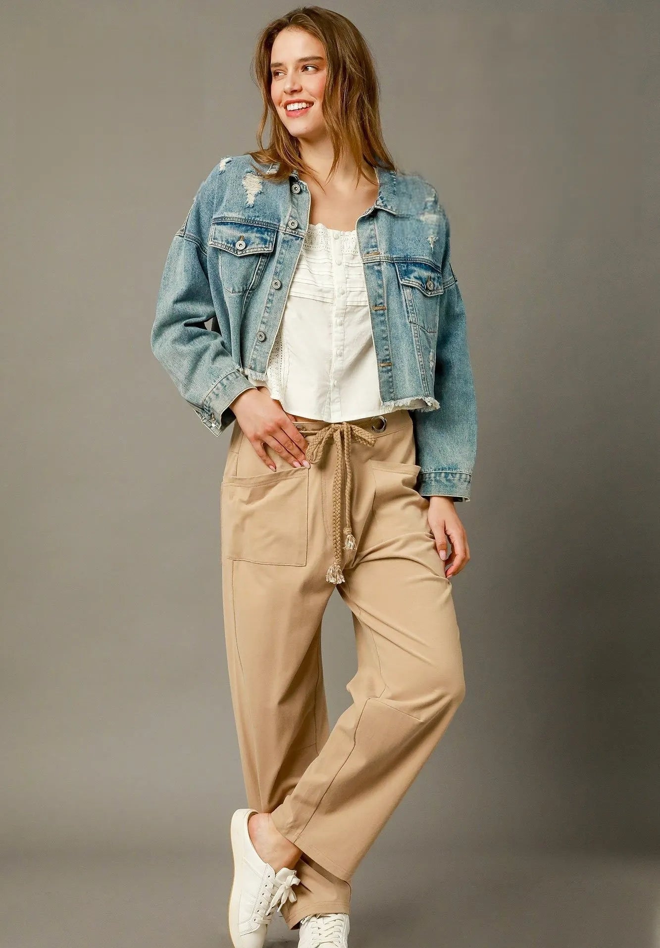 Woman wearing a denim jacket, white shirt, beige pants, and white sneakers on a gray background