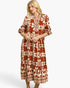 A woman modeling a rust-orange and cream patterned midi dress with three-quarter sleeves and a gathered waist, set against a plain light gray studio background.