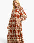 A woman modeling a long-sleeved maxi dress with a rust-orange and cream bohemian floral pattern against a light gray studio background.