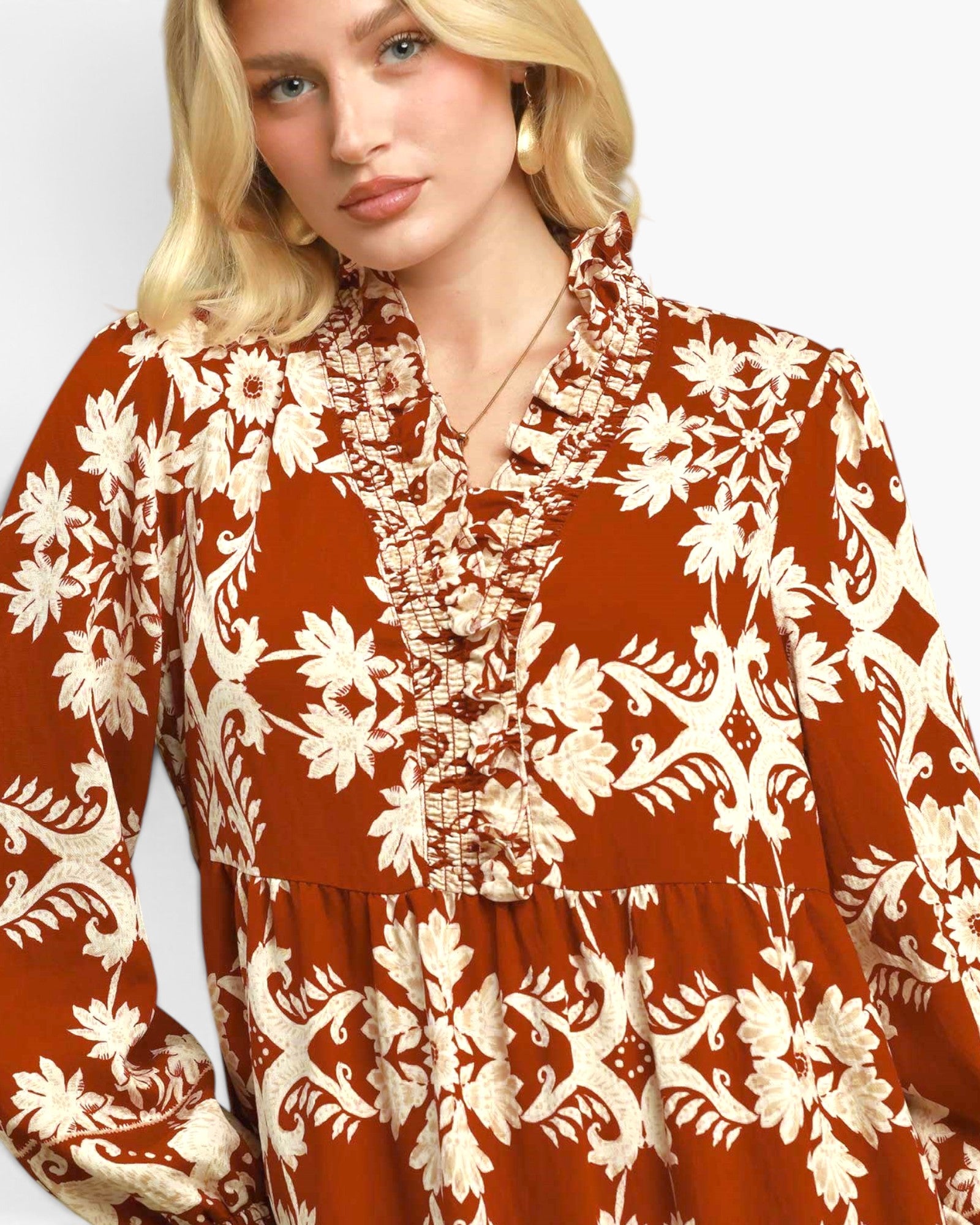 A close-up of a woman wearing a rust-orange long-sleeve blouse with a cream floral and scroll botanical pattern, featuring a ruffled high collar and buttoned front placket.
