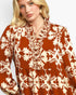 A close-up of a woman wearing a rust-orange long-sleeve blouse with a cream floral and scroll botanical pattern, featuring a ruffled high collar and buttoned front placket.
