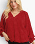 A woman wearing a red crinkled blouse with a V-neckline and voluminous long sleeves, paired with black bottoms, set against a plain light gray background.