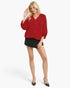 A blonde model wearing a red long-sleeve V-neck blouse with balloon sleeves, a black mini skirt, and black pointed-toe mule heels, standing against a light gray studio background.