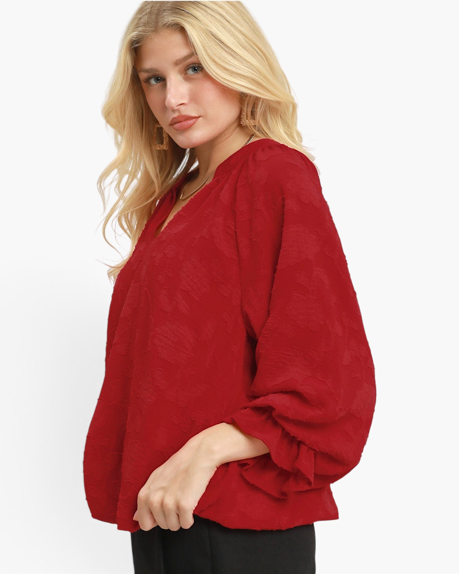 A blonde woman wearing a red textured long-sleeve blouse with a V-neck and black pants, photographed against a clean white studio background.