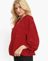 A blonde woman wearing a red textured long-sleeve blouse with a V-neck and black pants, photographed against a clean white studio background.