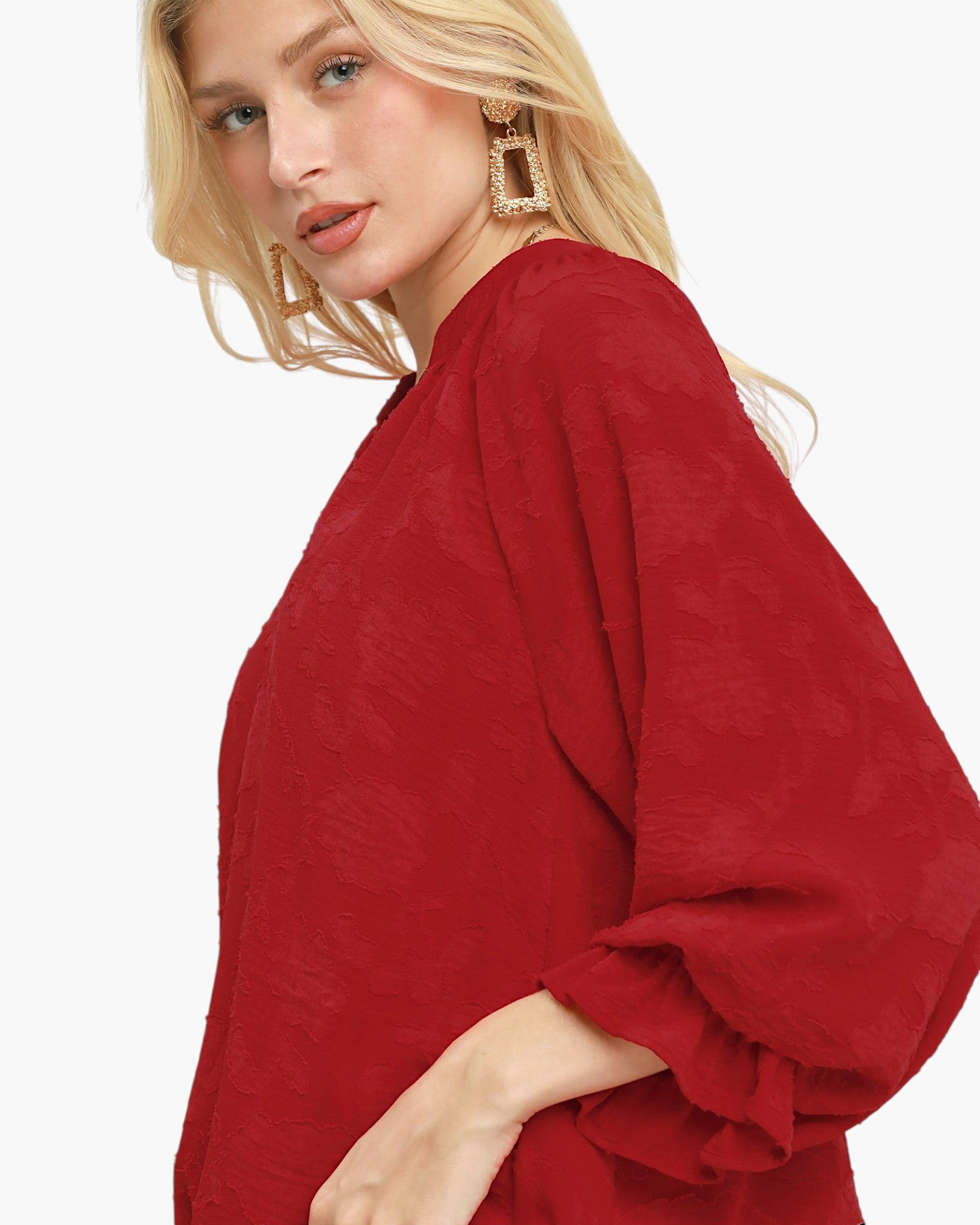 A close-up of a blonde woman wearing a textured red long-sleeve blouse with a high neckline and voluminous sleeves, set against a plain white background.