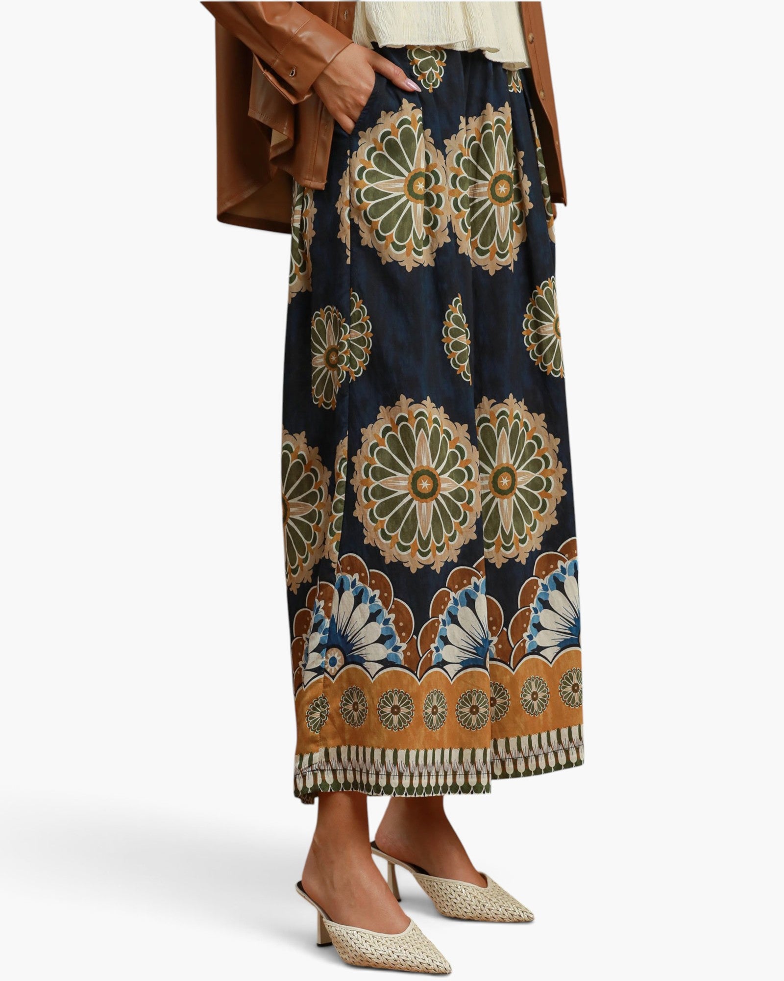 A model wearing a navy blue maxi skirt with a large, intricate mandala and floral pattern in beige, brown, and blue. The skirt features a decorative scalloped border and is styled with a brown jacket and beige pointed-toe mule heels against a clean white background.