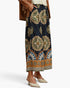 A model wearing a navy blue maxi skirt with a large, intricate mandala and floral pattern in beige, brown, and blue. The skirt features a decorative scalloped border and is styled with a brown jacket and beige pointed-toe mule heels against a clean white background.