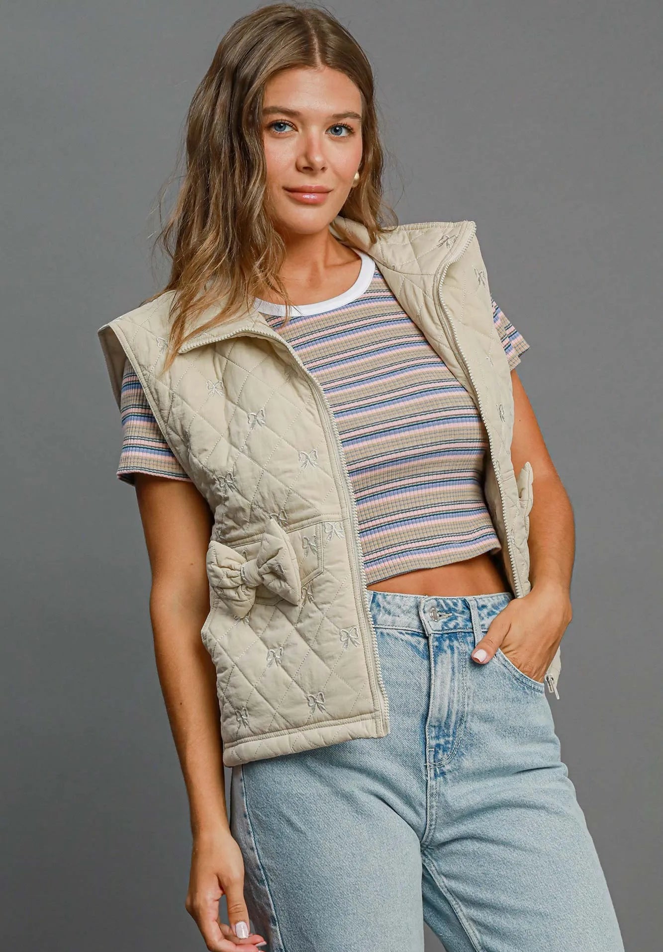 Woman wearing a beige quilted vest over a striped shirt with light blue jeans on a gray background
