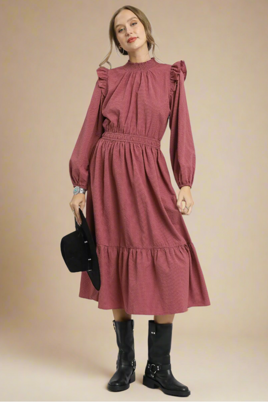 Woman wearing a dusty rose dress with ruffled sleeves and waistband, holding a black handbag on a beige background.