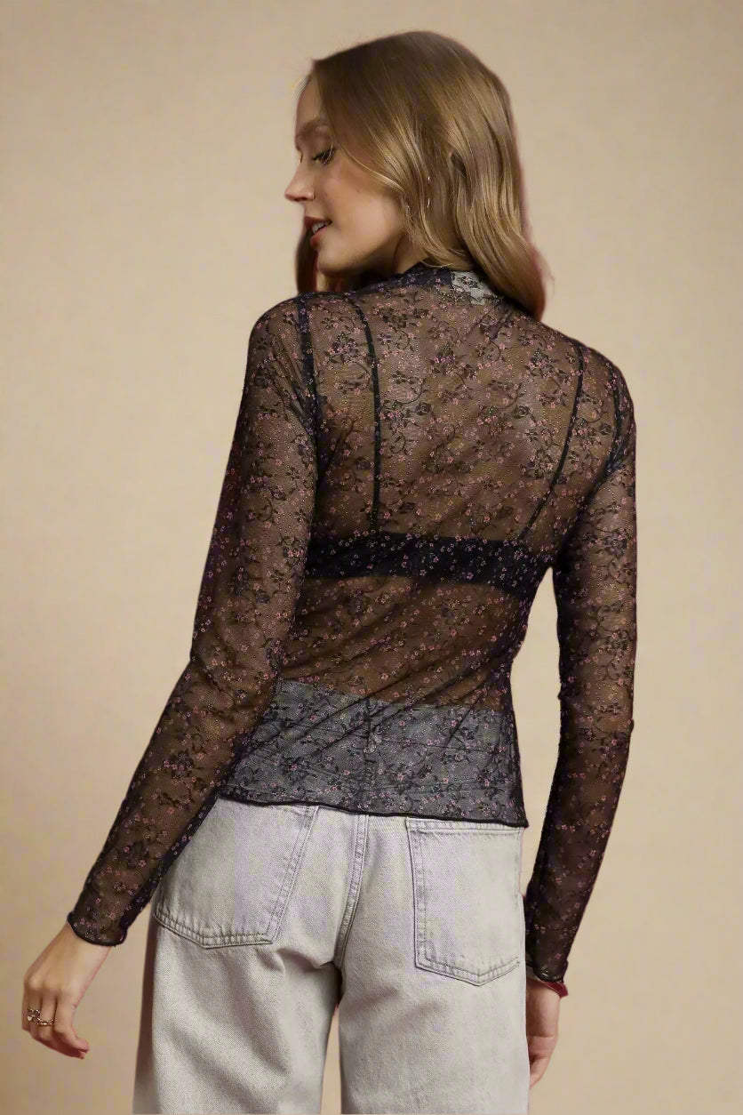 Woman wearing a sheer black lace top over a gray shirt with light gray pants on a red background.