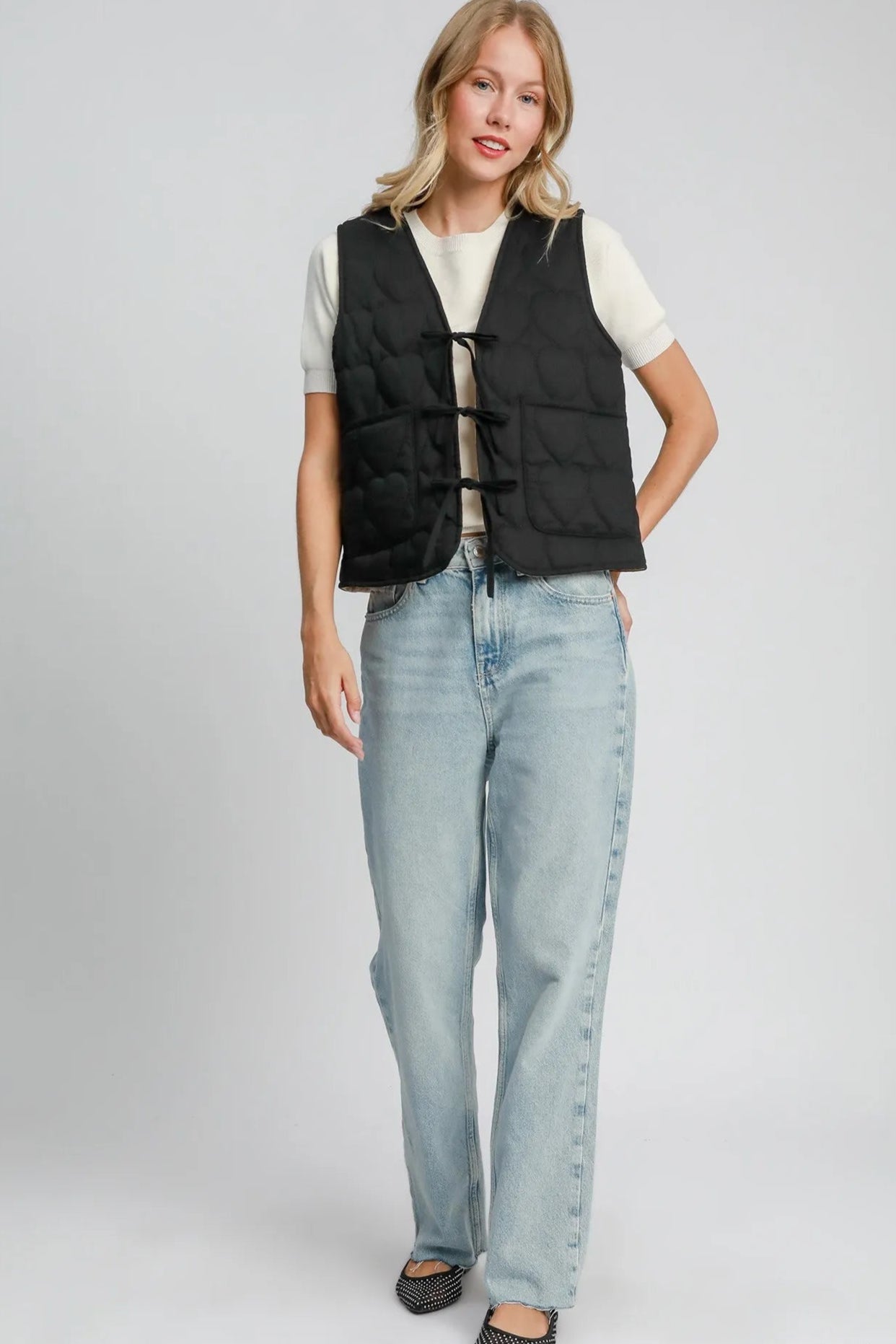 Woman wearing a black vest, white shirt, and light blue jeans on a gray background