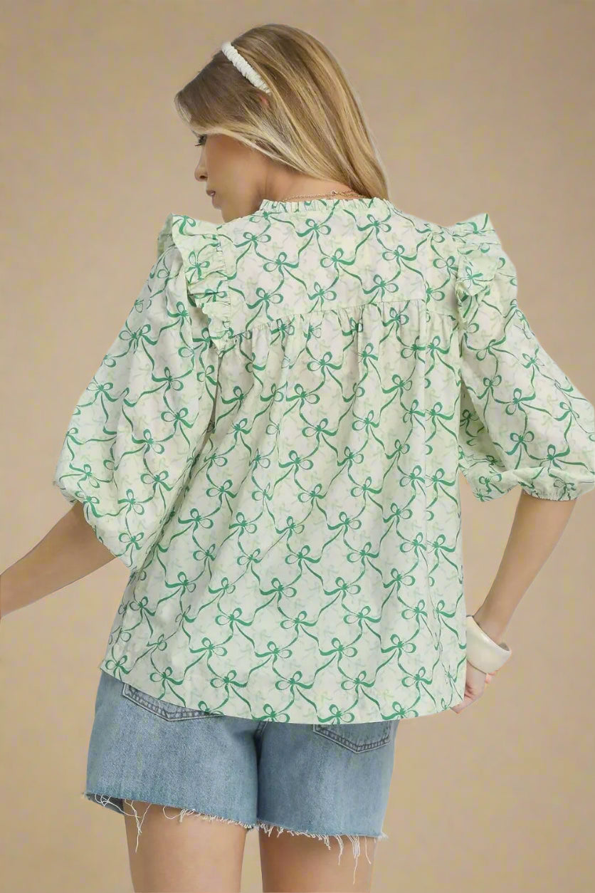 Woman wearing a light green blouse with a pattern of small shapes, paired with denim shorts on a plain background.