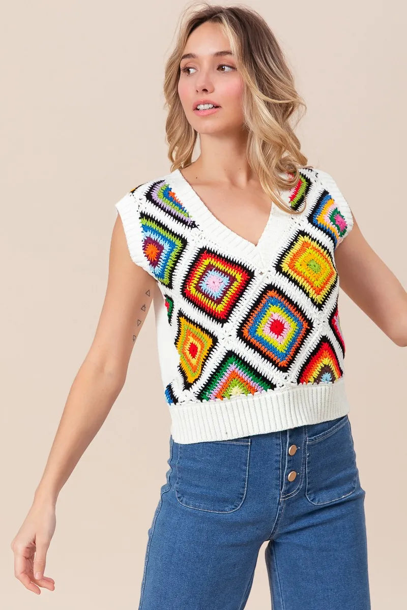 Woman wearing a colorful crochet vest over a white shirt with blue jeans on a beige background