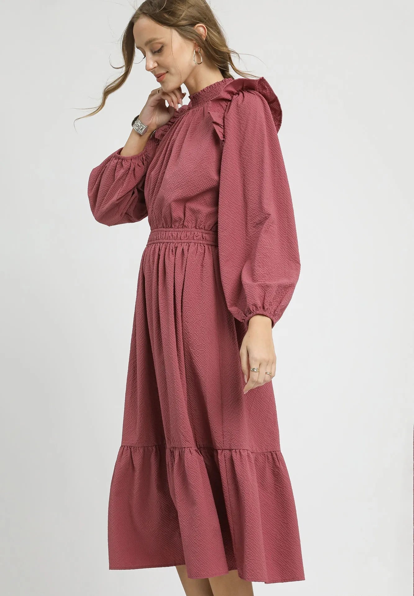 Woman wearing a dusty rose dress with long sleeves and ruffled details on a white background.