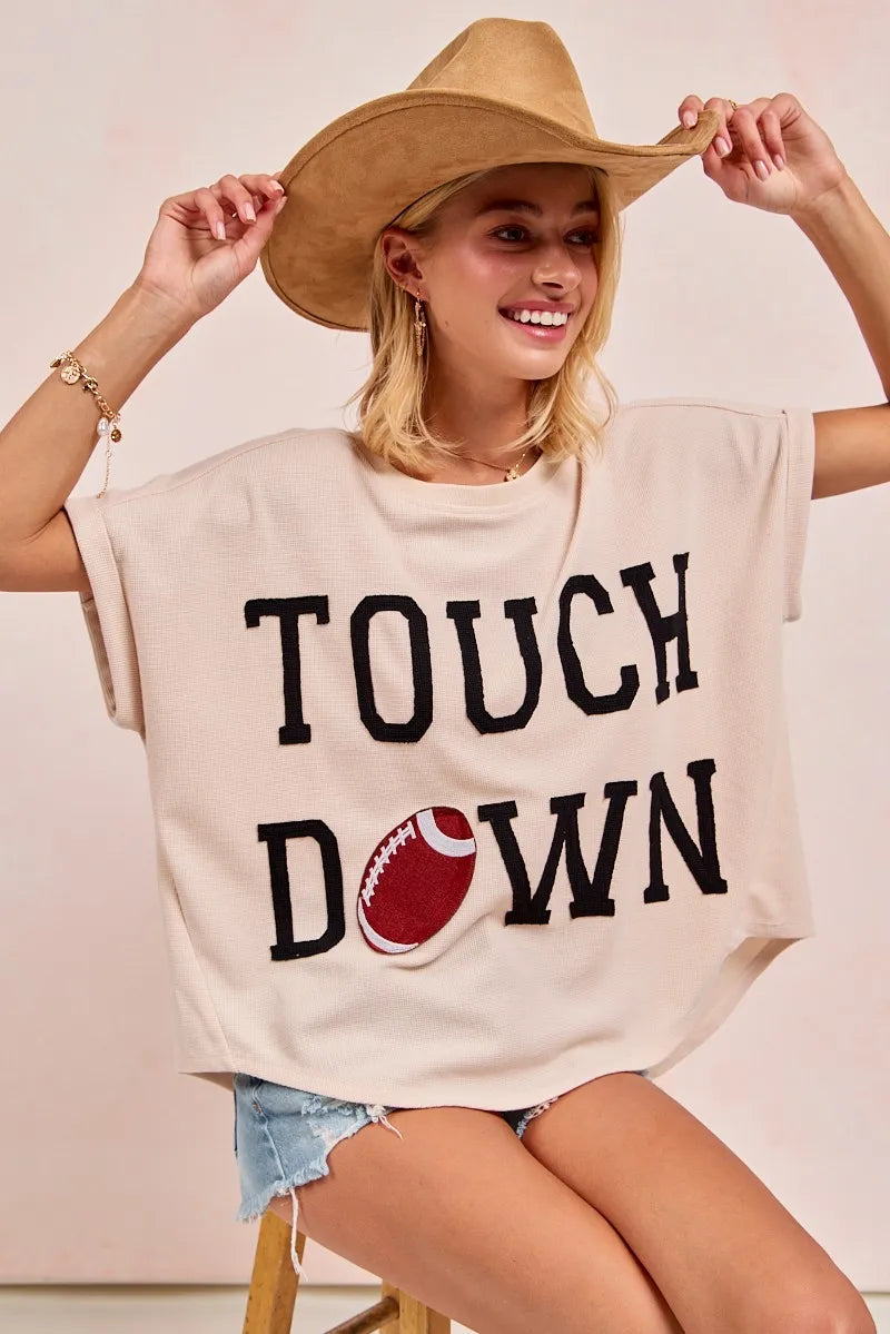 Woman wearing a beige oversized t-shirt with 'TOUCHDOWN' text and football graphic, holding a cowboy hat.