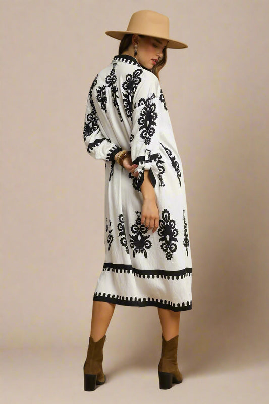 Woman wearing a white dress with black patterns, brown hat, and boots on a beige background