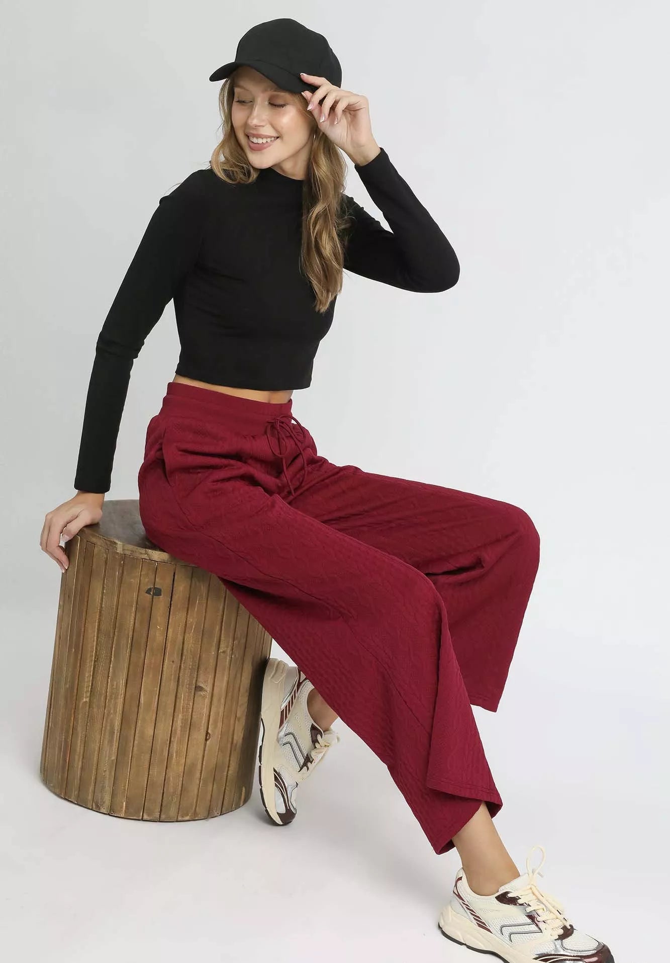 Woman sitting on a wooden stool wearing a black top and red pants, with a white background.