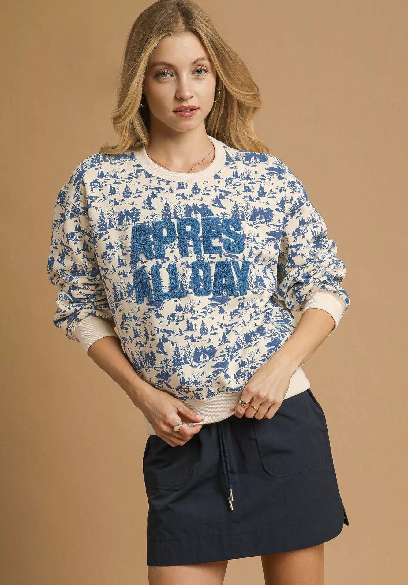Woman wearing a patterned sweater with 'APRES ALLDAY' text and a navy skirt on a beige background