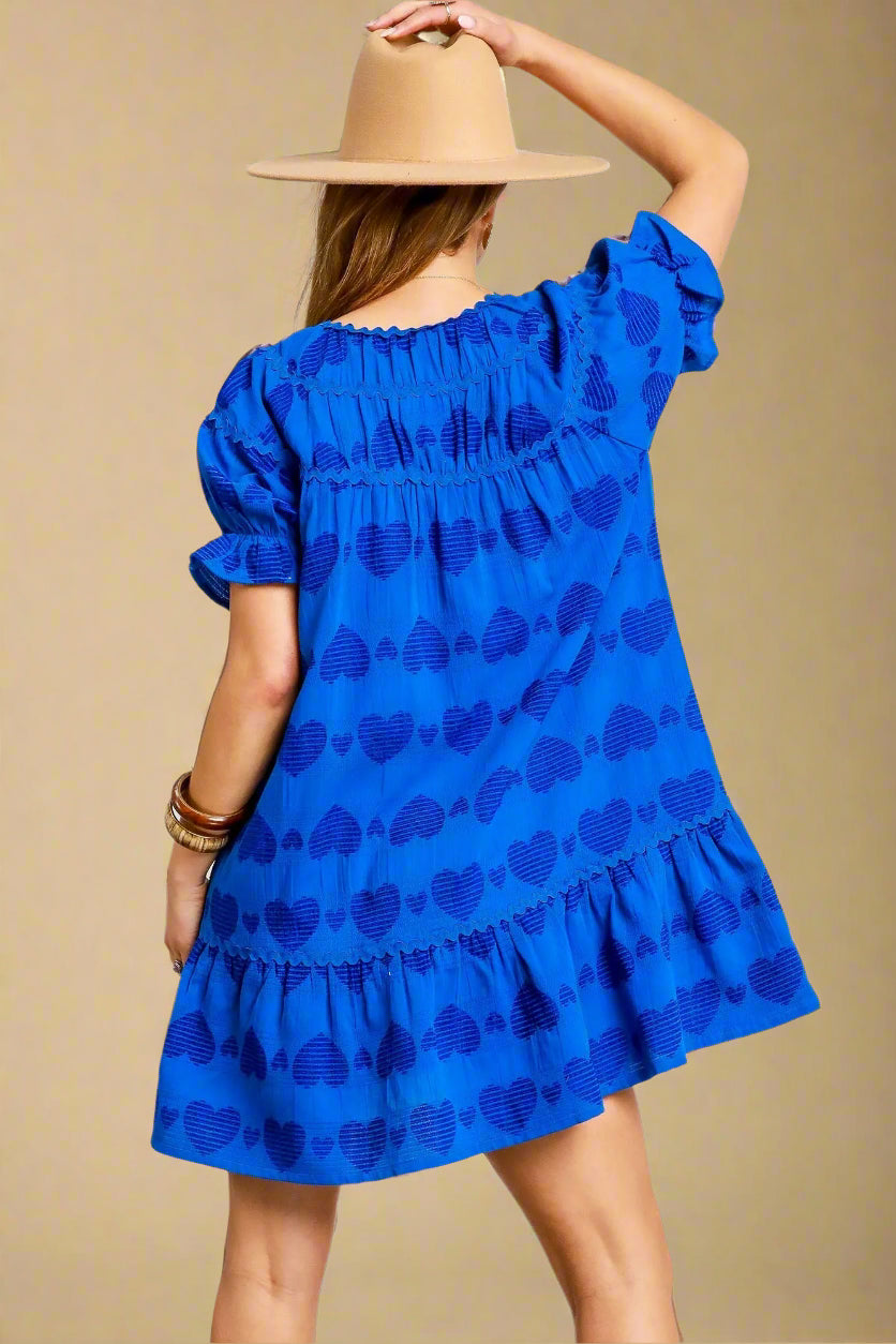 Woman wearing a blue dress with a pattern, holding a beige hat, on a light gray background.