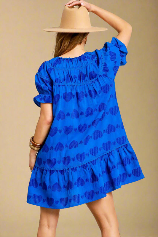 Woman wearing a blue dress with a pattern, holding a beige hat, on a light gray background.