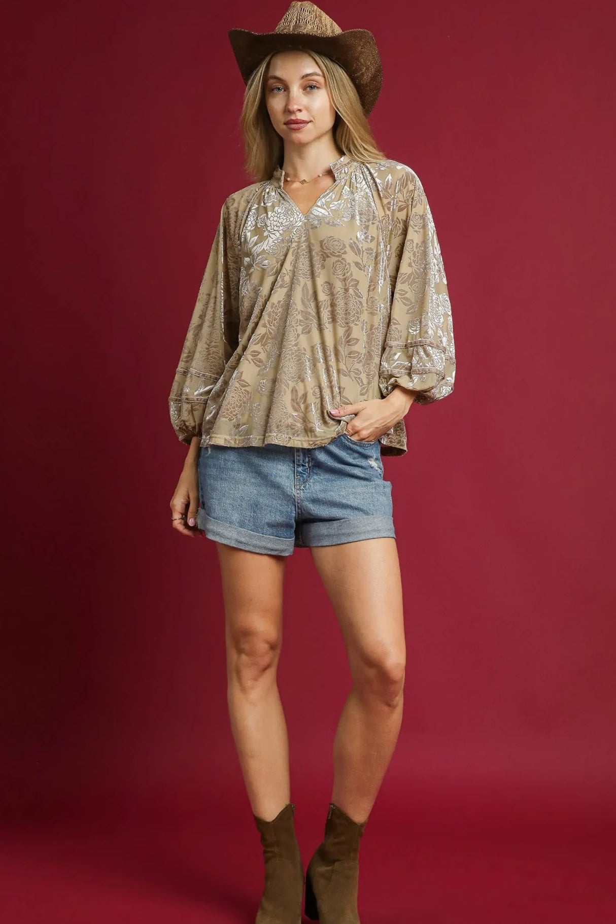 Woman wearing a patterned blouse, denim shorts, and cowboy hat against a red background