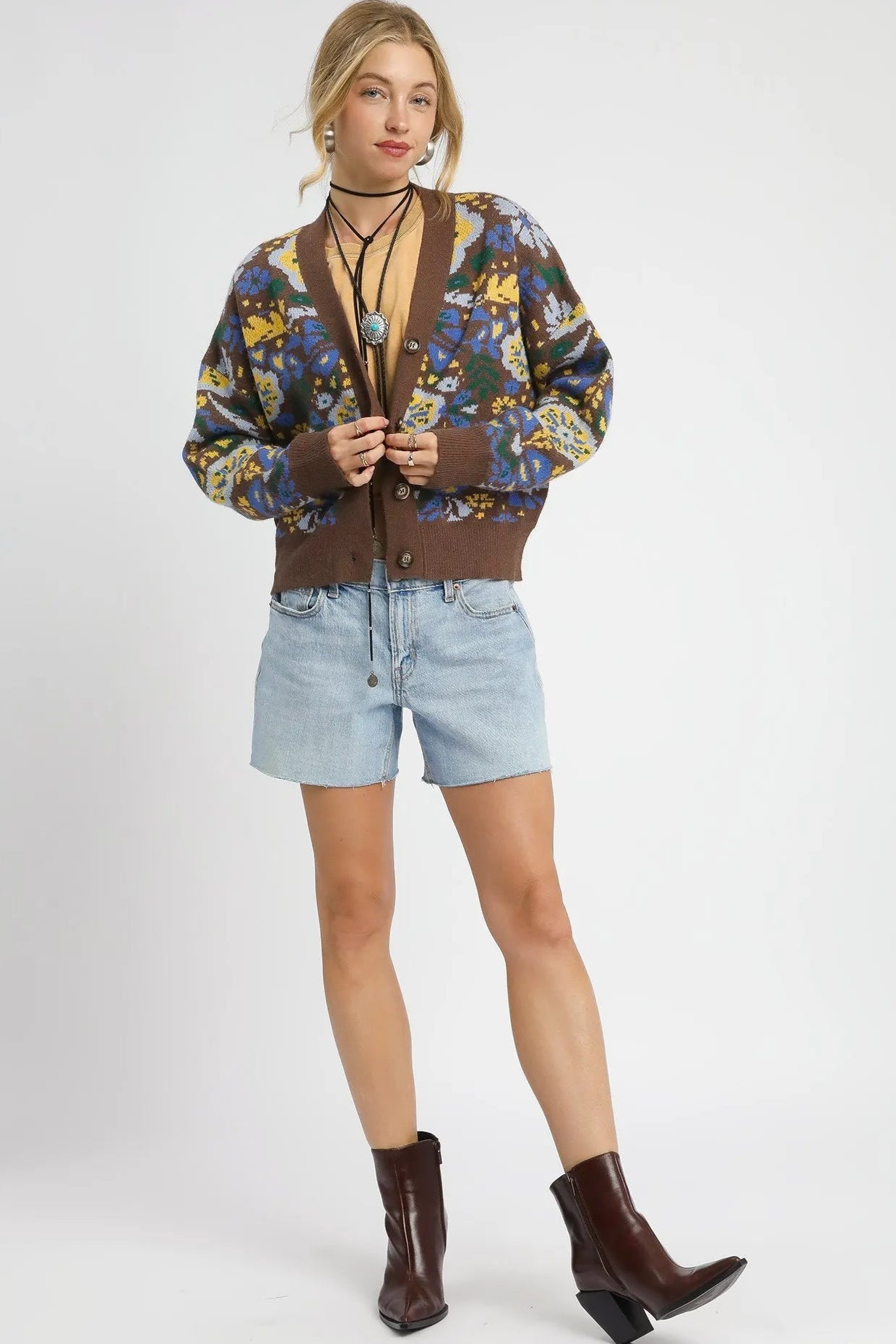 Woman wearing a patterned cardigan, denim shorts, and brown boots on a white background