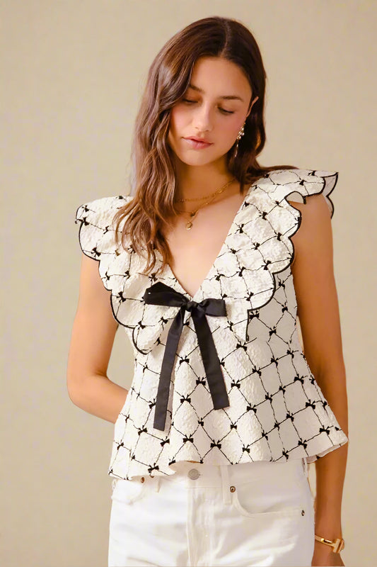 Woman wearing a black and white patterned blouse with a bow tie, standing in front of a wooden door and cactus plant.