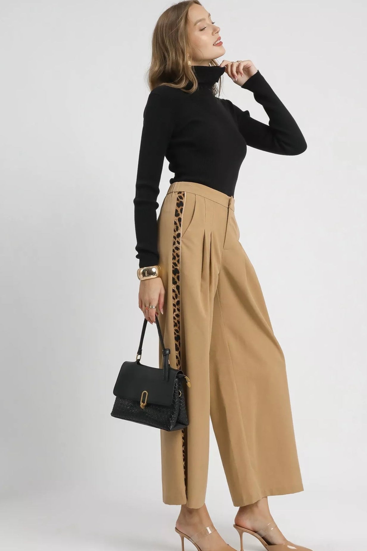 Woman wearing a black turtleneck and beige pants with leopard print details, holding a black handbag on a white background.
