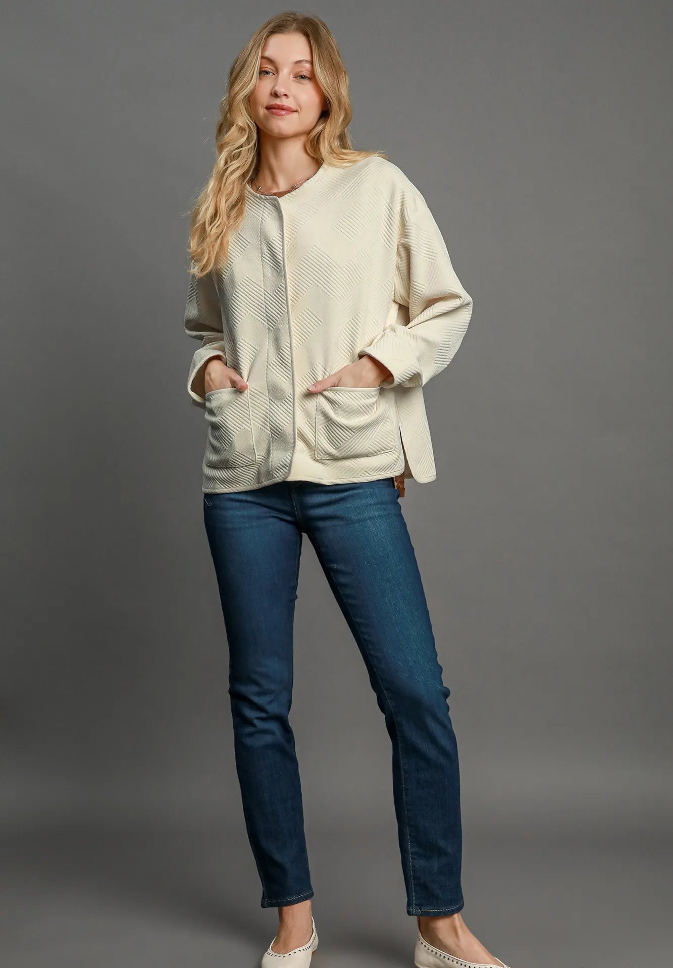 Woman wearing a cream cardigan and blue jeans against a gray background