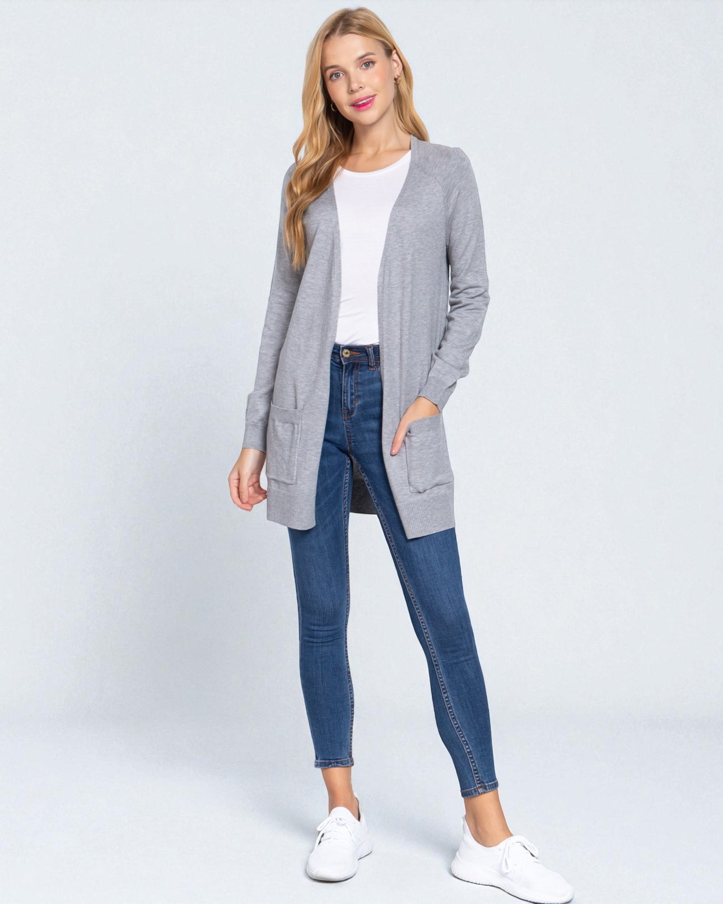 Woman wearing a gray cardigan, white shirt, blue jeans, and white sneakers on a white background