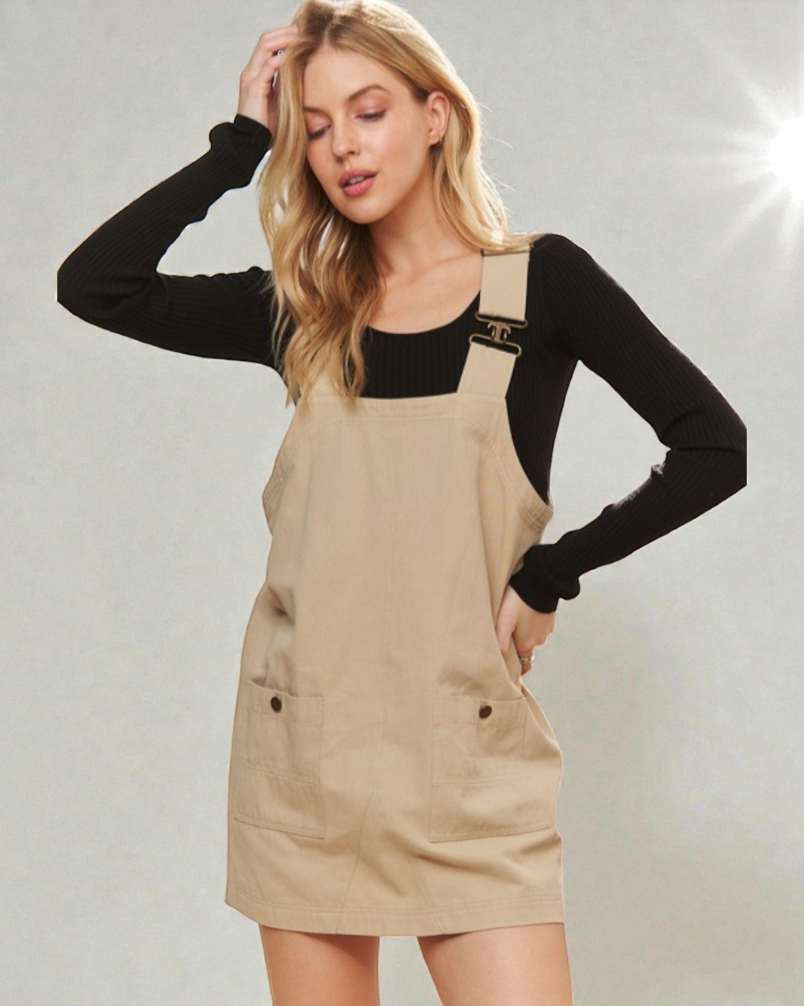 Woman wearing a beige pinafore dress over a black long-sleeve shirt on a gray background