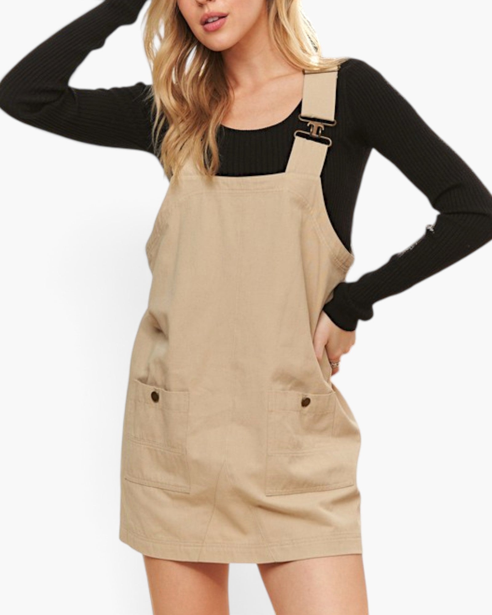 A woman wearing a beige corduroy bib overalls dress over a black long-sleeve top, shown from the chest down against a plain white background.