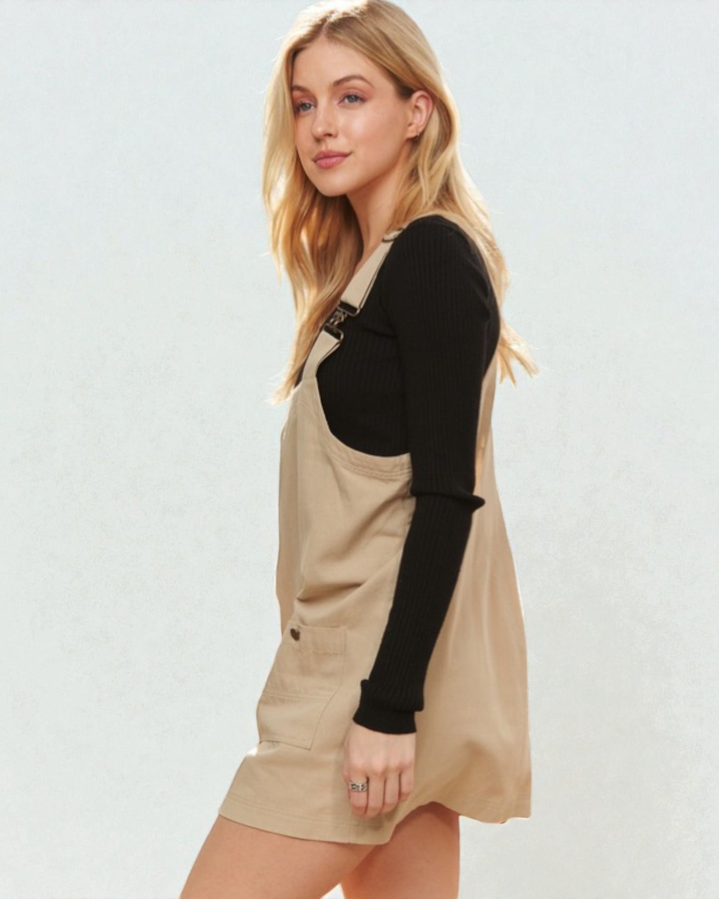 Woman wearing a beige pinafore dress over a black long-sleeve shirt on a white background