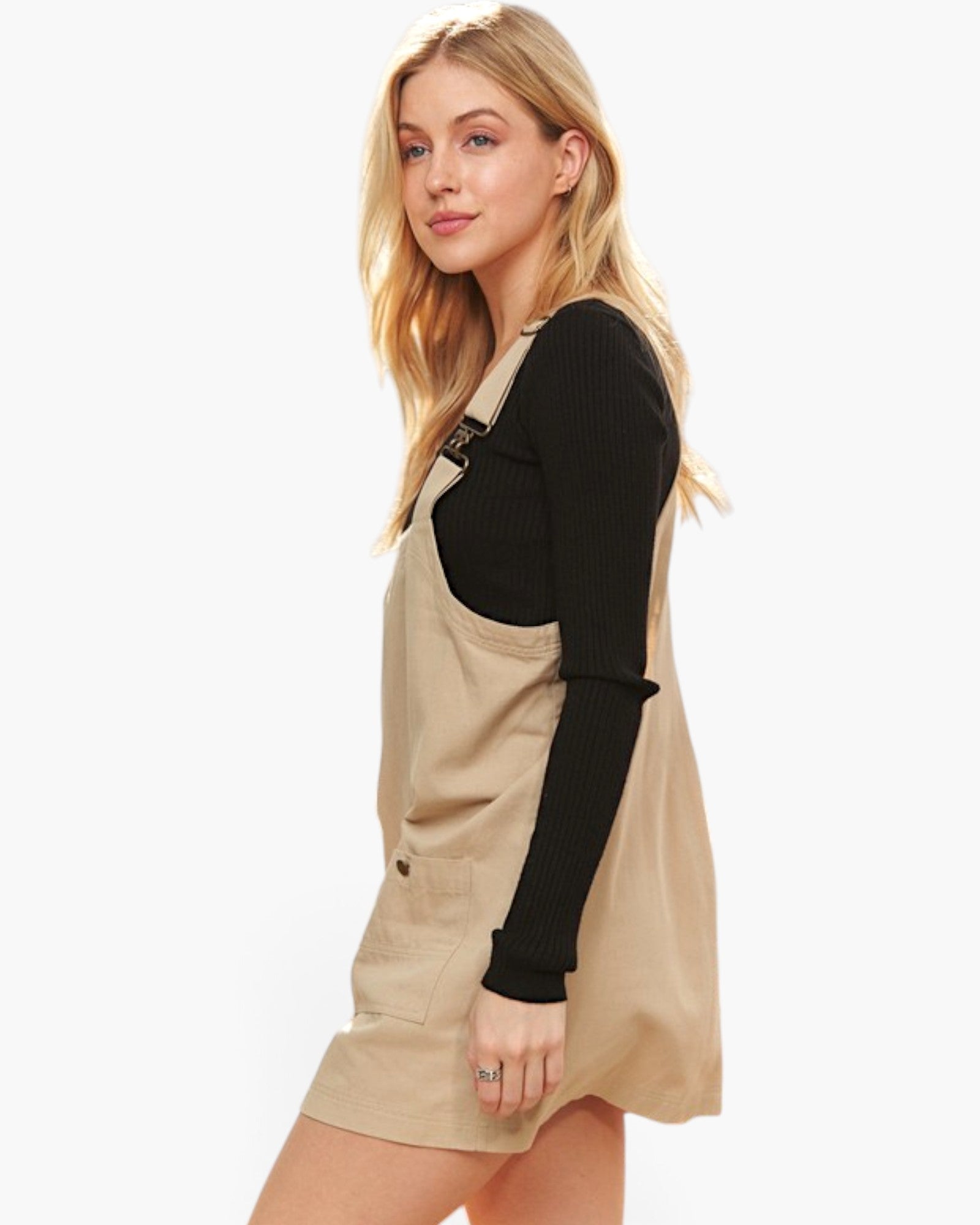 A blonde woman modeling a beige overall mini dress over a black ribbed long-sleeve top, photographed against a clean white background.